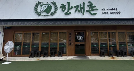 가게 앞 — 흰 간판에 초록 소 로고와 «한재촌», «청도 미나리마을 · 소찌개, 육회비빔밥», 나무 창틀 가게 앞과 인조잔디 앞마당